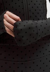 Chaqueta atlética negra con textura de puntos, cierre de cremallera y orificios para el pulgar. La tela es suave con un ligero brillo.