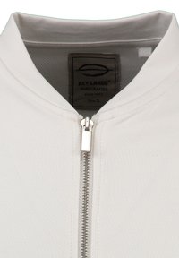 Primer plano del cuello de una chaqueta blanca con una cremallera plateada central y una etiqueta interna visible que dice "KEY LARGO HECHO A MANO Talla S."