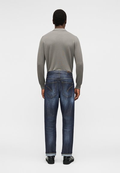 Άνδρας που φοράει γκρι μακρυμάνικο πουκάμισο, μπλε τζιν με γυρισμένα ρεβέρ και μαύρα παπούτσια, στέκεται απέναντι από ένα απλό ανοιχτό γκρι φόντο.