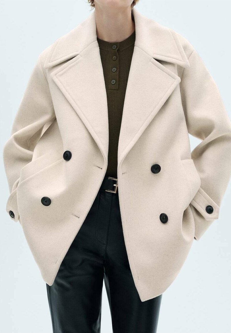Femme portant un manteau en laine beige à double boutonnage sur un pull vert foncé boutonné et un pantalon en cuir noir avec une ceinture noire.