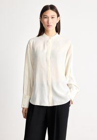 Krém színű, hosszú ujjú blúz mandarin gallérral és finom fényű felülettel. Gombos záródással és széles mandzsettákkal rendelkezik; laza szabás a kényelemért.