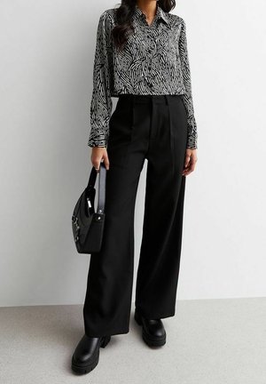 Persoon draagt zwarte broek met wijde pijpen, zwarte grove laarzen, een patroonrijke lange mouwen blouse en houdt een zwarte handtas naast zich vast.