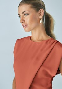Hliněné šaty s hladkou texturou, s kulatým výstřihem a draped detaily na rameni. Zlaté kruhové náušnice zvýrazňují styl.