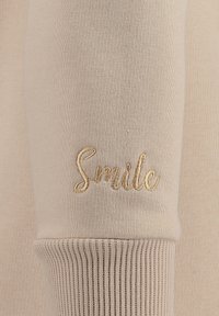 Maglia beige a maniche lunghe con polsini a coste; presenta la parola ricamata "Smile" in filo dorato sulla manica. Tessuto morbido e liscio.