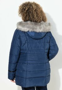 Mörkblå puffjacka med fodrad huva som har fuskpälsbrodyr, quiltat mönster och en något längre rygg för extra värme.