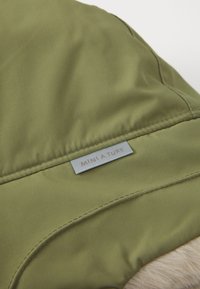 Tissu matelassé vert olive avec des coutures piquées et une petite étiquette grise portant l'inscription "MINI A TURE," associé à une bordure en fausse fourrure tan texturée.