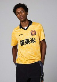 Camiseta de fútbol amarilla con acentos negros, cuello y gran texto en negro. Presenta un diseño con patrón y un logotipo en la zona del pecho izquierdo.