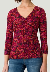 Femme portant un haut ajusté à manches longues avec un col en V torsadé et un motif floral paisley vibrant rouge, rose et orange, associé à un jean bleu.