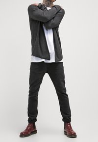 Grijze lange mouwen cardigan over een wit T-shirt, gecombineerd met zwarte broek en bordeauxrode enkellaarsjes. Casual fit en modern design.