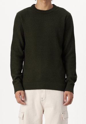 Homme portant un pull en maille texturée vert foncé et un pantalon cargo beige clair, debout devant un fond blanc.
