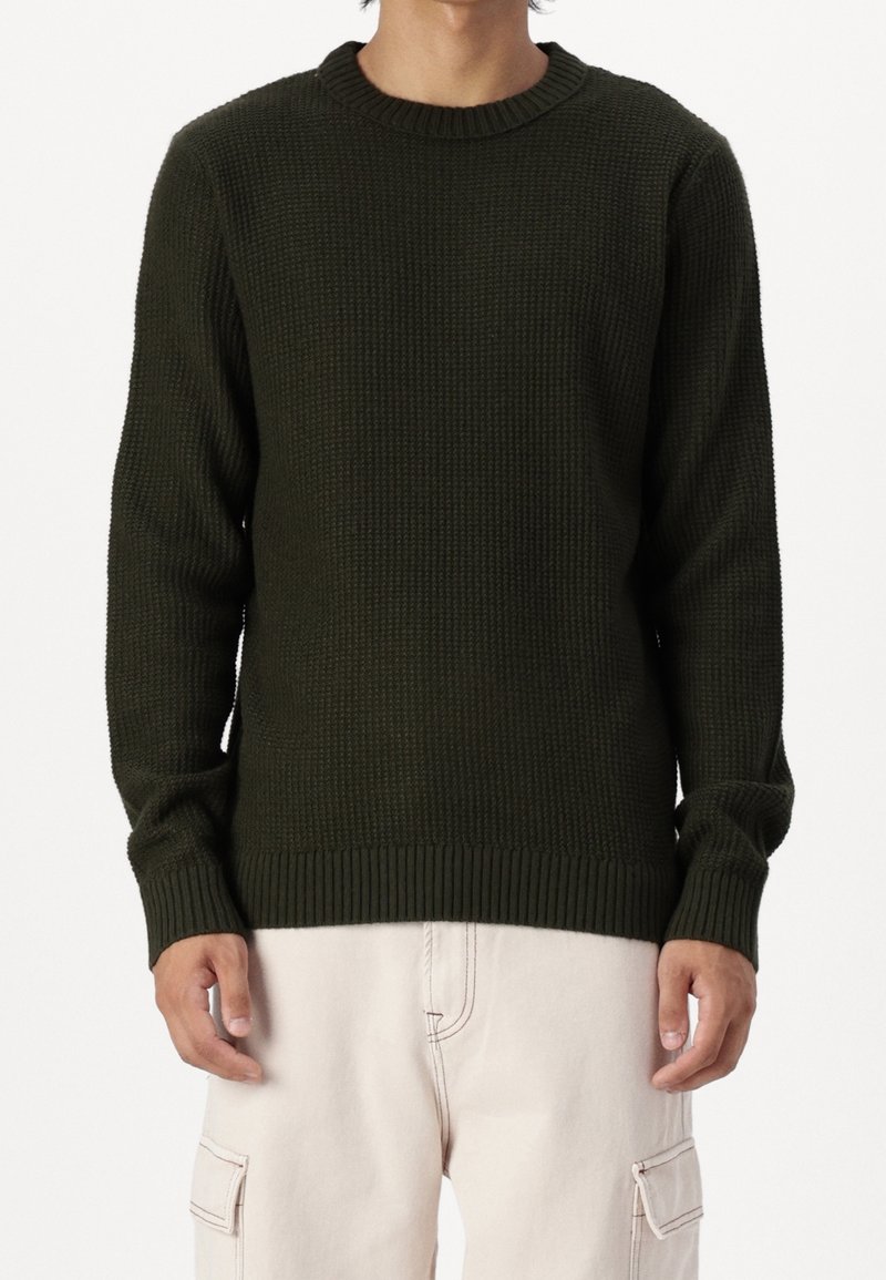 Homme portant un pull en maille texturée vert foncé et un pantalon cargo beige clair, debout devant un fond blanc.