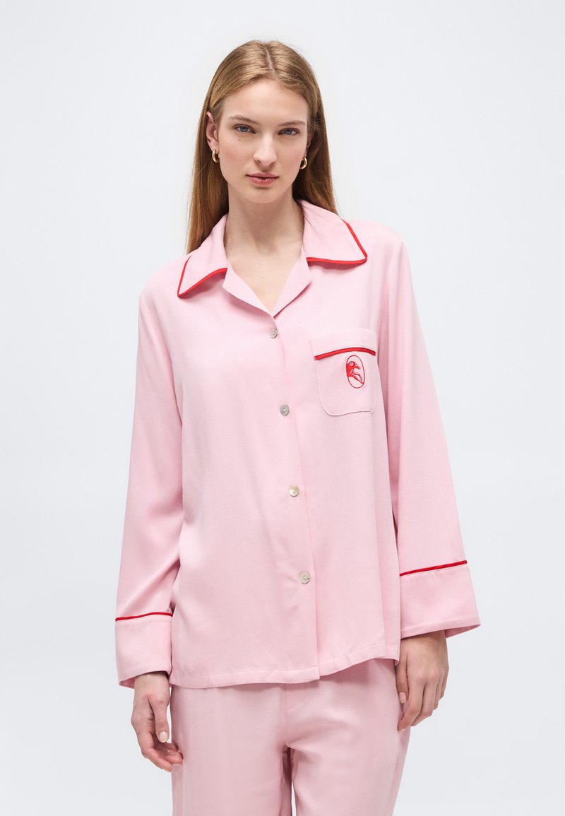 Γυναίκα που φοράει ανοιχτό ροζ σατέν πιτζάμα με κόκκινη επένδυση και μικρό κεντητό κόκκινο φλαμίνγκο στην τσέπη στο στήθος, στέκεται μπροστά σε λευκό φόντο.