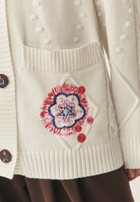 Biały dziergany kardigan z fakturowanymi pomponami i prążkowanymi brzegami. Ozdobiony kolorowym haftem kwiatowym na lewym kieszeni, zapinany na guziki.