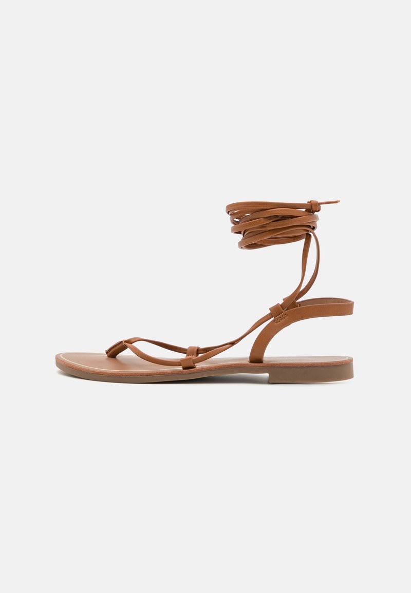 Sandal de couro castanho com sola plana, apresentando tiras finas e um envoltório em espiral para o tornozelo. Textura suave e design minimalista.