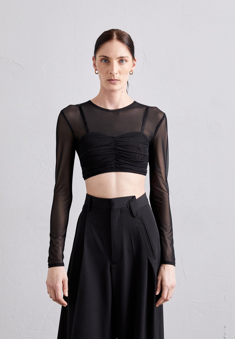 Svart cropped topp med lange gjennomsiktige ermer og rynkedetaljer ved brystet, parret med høyt liv svarte bukser med folder.
