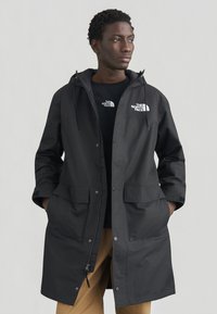 Μαύρο αδιάβροχο παρκά με κουκούλα, διαθέτει μπροστινά κουμπώματα με τρουκ, δύο μεγάλες τσέπες και λογότυπο στην αριστερή πλευρά του στήθους.