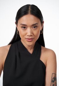 Top nero con scollo a halter e dettagli plissettati, orecchini a cerchio dorati tondi e capelli lisci scuri, su uno sfondo bianco uniforme.