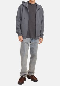 Haut à capuche zippé gris avec un cordon de serrage, associé à un t-shirt sombre, un jean bleu clair et des chaussures à lacets marron.