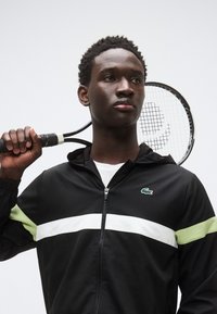 Musta vetoketjullinen urheilutakki, jossa valkoiset ja vaaleanvihreät raidat, rakenteellinen kangas, yhdistetty tennismailaan. Lacosten logo vasemmassa rinnassa.
