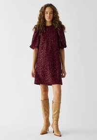 Rochie mini din sequins burgundy, cu mâneci bufante și guler rotund, asortată cu cizme bej cu toc stiletto până la genunchi.