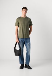 Moški stoji v zeleni kratkotrajni srajci, modrih kavbojkah, črnih supergah, drži majhno črno torbico z logotipom "BOSS" pred enobarvno belo ozadje.