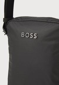 Černá polstrovaná ramenní taška s hladkým povrchem, na kterém se nachází lesklé logo "BOSS". Obsahuje zipové zapínání a nastavitelný popruh.