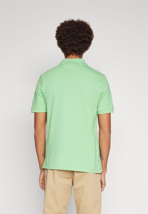 Man met krullend haar die een lichtgroen poloshirt met korte mouwen en beige broek draagt, met zijn rug naar een effen achtergrond toe.
