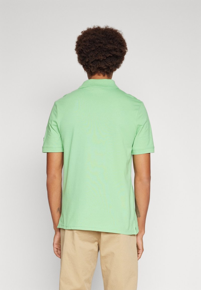 Homme aux cheveux bouclés portant un polo vert clair à manches courtes et un pantalon beige, tourné de dos devant un fond uni.