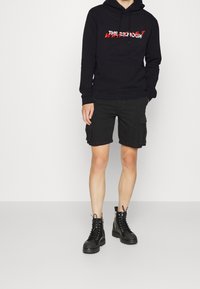Sudadera negra con logo frontal, pantalones cortos cargo negros y botas de tobillo negras con cordones. El material parece suave, con un corte relajado y bolsillos.