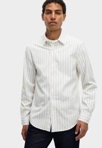 Camisa blanca de botones con finas rayas verticales en gris claro y rojo oscuro. Presenta mangas largas y un cuello clásico. Combinada con jeans oscuros.
