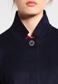 Marinblå ullkappa med hög krage och en enkel svart knappstängning, med en slät textur och minimala designelement.