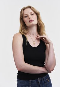 Woman with long red hair wearing a black sleeveless top, blue jeans, and a gold pendant necklace, posing with one hand near her neck.