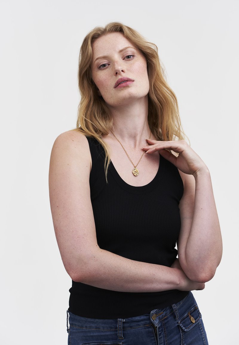 Woman with long red hair wearing a black sleeveless top, blue jeans, and a gold pendant necklace, posing with one hand near her neck.