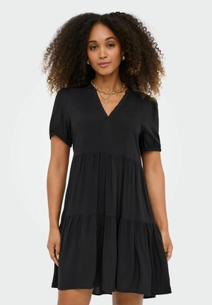 Ženska s kodrastimi lasmi, oblečena v kratkoročno črno obleko z več plastmi in z zlatim verižico, stoji pred enobarvnim svetlim ozadjem.