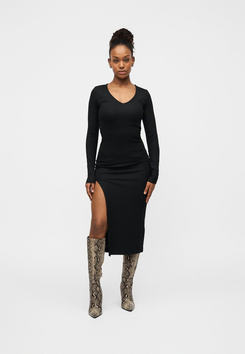 Μαύρο μακρυμάνικο φόρεμα με V-λαιμόκοψη και άνοιγμα στο πλάι, συνδυασμένο με μπότες έως το γόνατο με σχέδιο φίδι. Ομαλή υφή υφάσματος.