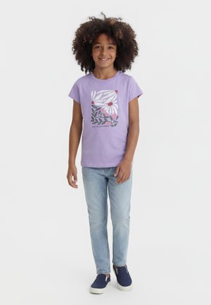 Uśmiechnięte dziecko z kręconymi włosami, ubrane w lawendowy t-shirt z kwiatowym nadrukiem, jasnoniebieskie dżinsy i granatowe wsuwane buty, stojące na białym tle.