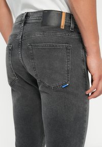 Πίσω όψη ατόμου που φοράει γκρι τζιν παντελόνι με ραμμένα τσέπες και ένα μαύρο δερμάτινο λογότυπο στη μέση.