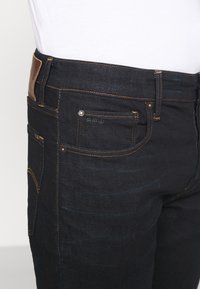 Jeans de mezclilla oscura con costuras naranjas, una parche de cuero en la cintura y múltiples bolsillos, incluido un pequeño bolsillo frontal para monedas.