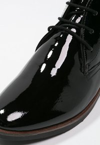 Zapato de charol negro con un acabado brillante, que presenta una punta redondeada, cordones y una suela marrón. Textura suave con un minimalismo en los detalles.
