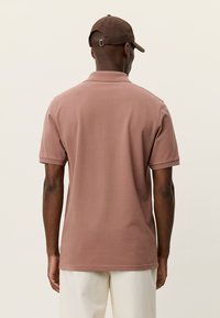 Καφέ πόλο μπλούζα με κοντά μανίκια και στρωτή γιακά. Μαλακό ύφασμα, μονόχρωμο και άνετη εφαρμογή. Φορεμένη με ανοιχτόχρωμο παντελόνι.