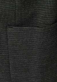 Strukturierter dunkelgrauer Stoff mit einem feinen Rautenmuster, der eine Taschenkonstruktion aufweist, und eine gewebte Struktur sowie einen subtilen Glanz demonstriert.