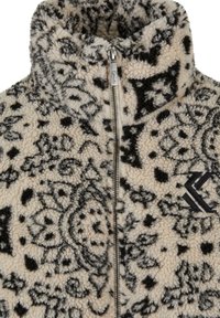 Fleece-takki, jossa on korkea kaulus ja etuvetoketju. Kangas on vaaleankermanvärinen, jossa on mustia koristeellisia kuvioita ja logo-yksityiskohta.