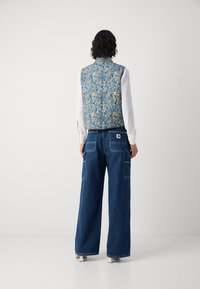 Γαλάζιο γιλέκο με ανθισμένα σχέδια και ψηλό γιακά, σε συνδυασμό με φαρδιά τζιν παντελόνια σε σκούρους τόνους που διαθέτουν πλαϊνές τσέπες και γυρισμένα μπατζάκια.