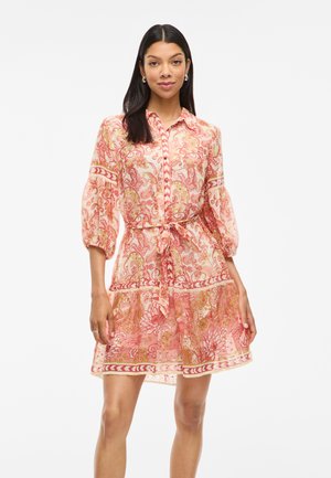 Femme portant une robe à motifs paisley rose, avec des manches bouffantes et une ceinture à la taille, se tenant devant un fond blanc uni.