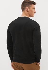 Svart långärmad tröja med rund halsringning, slät textur och ribbade muddar. Anmärkningsvärda detaljer inkluderar en enkel design och en söm på baksidan.