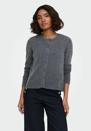 Femme aux cheveux courts et bouclés portant un cardigan gris boutonné et un pantalon taille haute foncé, debout devant un fond clair uni.