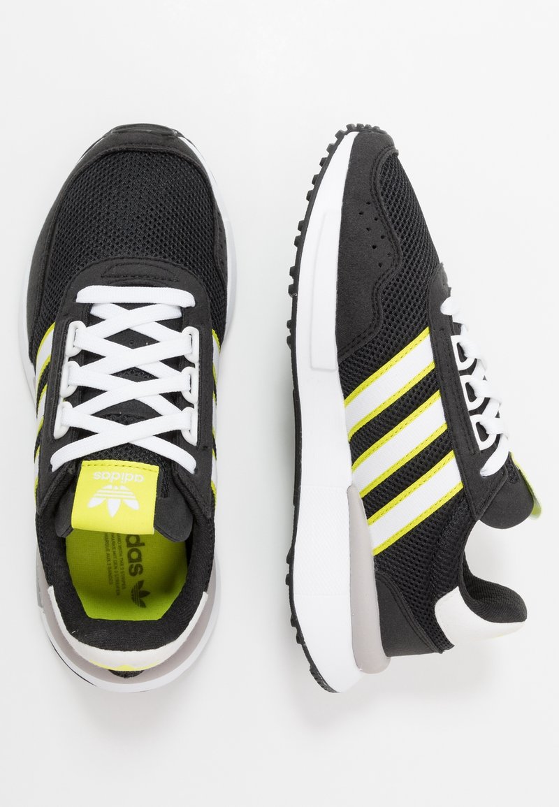 Chaussures de sport noires avec des lacets blancs, trois bandes blanches et des accents jaunes vifs. Confectionnées en mesh et en daim, avec une semelle amortie.