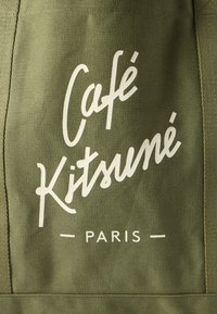 Olivazöld szövet, amelyen fehér szöveg olvasható: "Café Kitsuné" és "PARIS" modern írásmódban és sans-serif betűtípussal. A textúra sima.