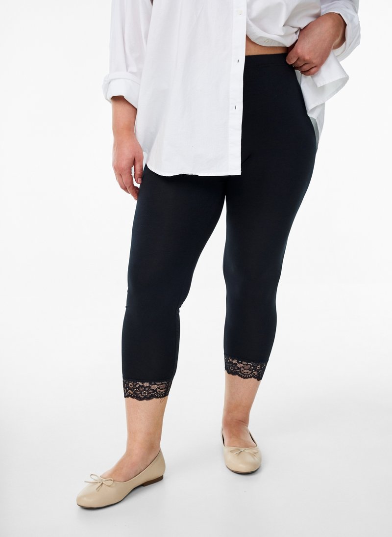 Persoon draagt zwarte cropped legging met kanten rand, beige ballerina's en een wit overhemd met oprolbare mouwen.