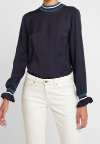 Blusa navy con maniche lunghe, colletto a coste blu e bianco, polsini arricciati, abbinata a pantaloni bianchi. Tessuto dalla texture liscia.